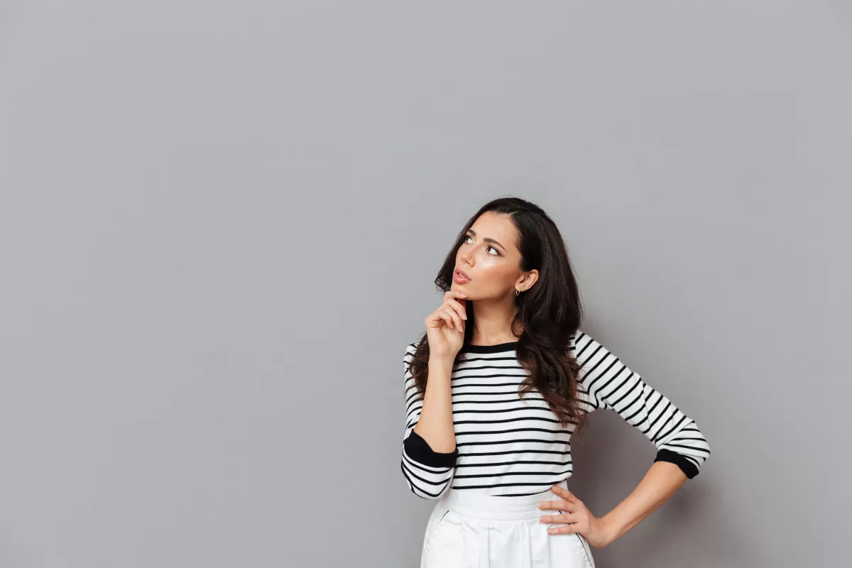 Woman in striped shirt and white skirt deep in thought against grey background.