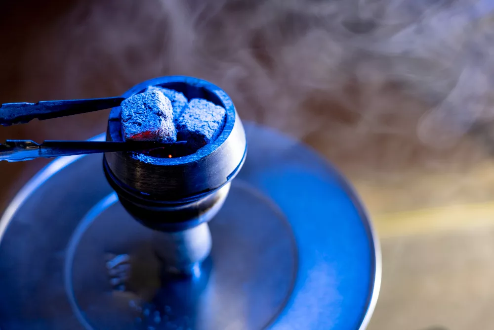 Glowing coal held by tongs above a hookah bowl with rising smoke.