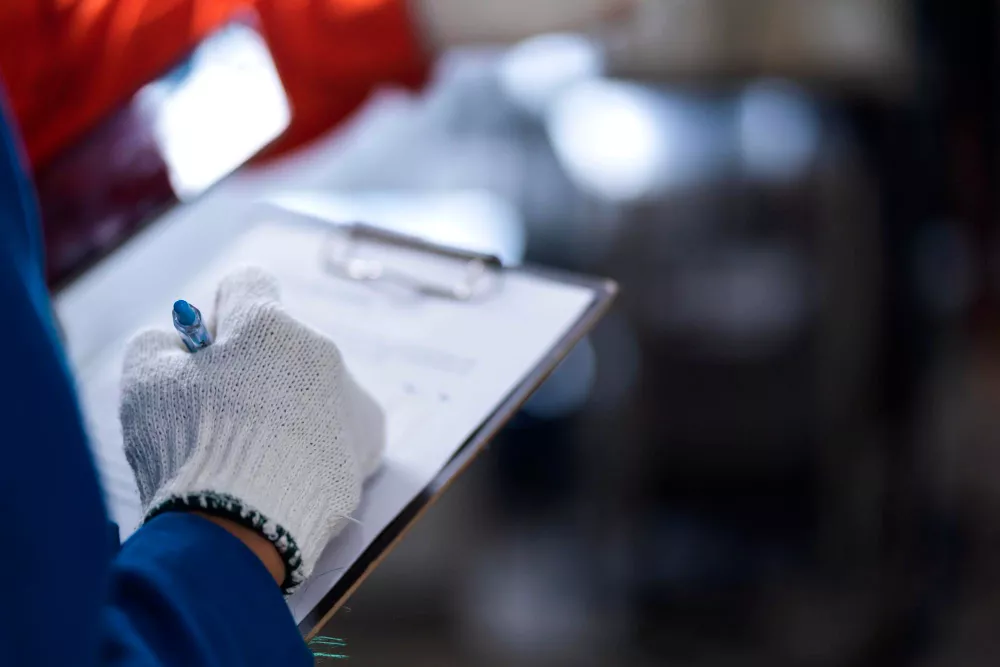 Worker in gloves holding pen and clipboard in industrial setting.