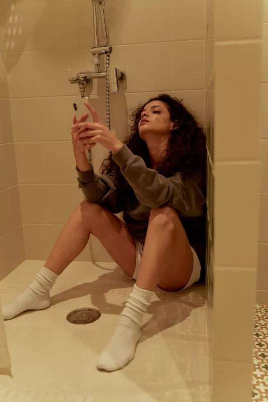Person sitting on shower floor, using smartphone in a cozy, intimate bathroom setting.
