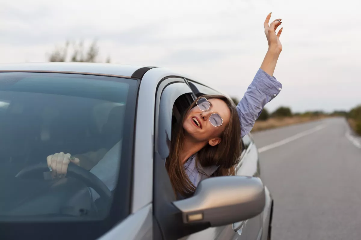 Woman enjoys freedom on road trip, arm raised in car, open road adventure ahead.