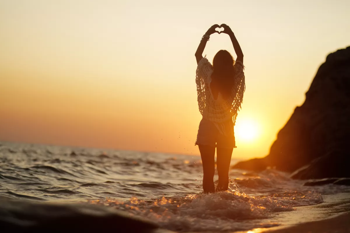 Woman forms heart silhouette against golden sunset by the sea.