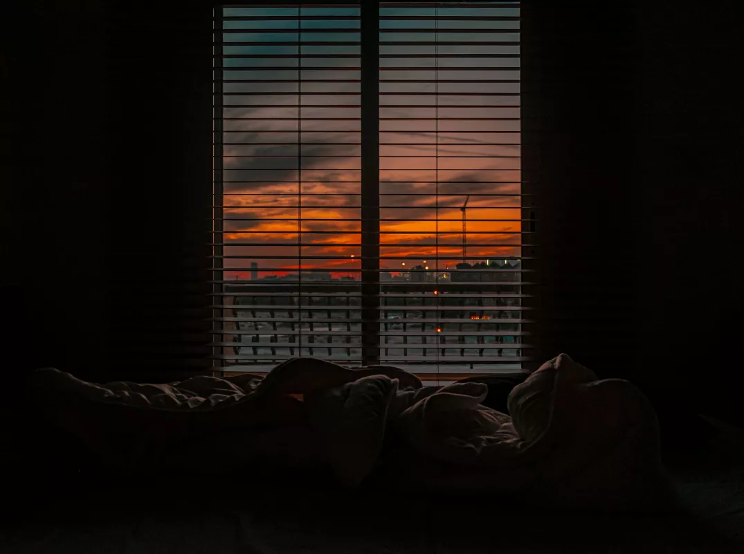 Serene bedroom with urban sunset view through blinds, peaceful city evening ambiance.