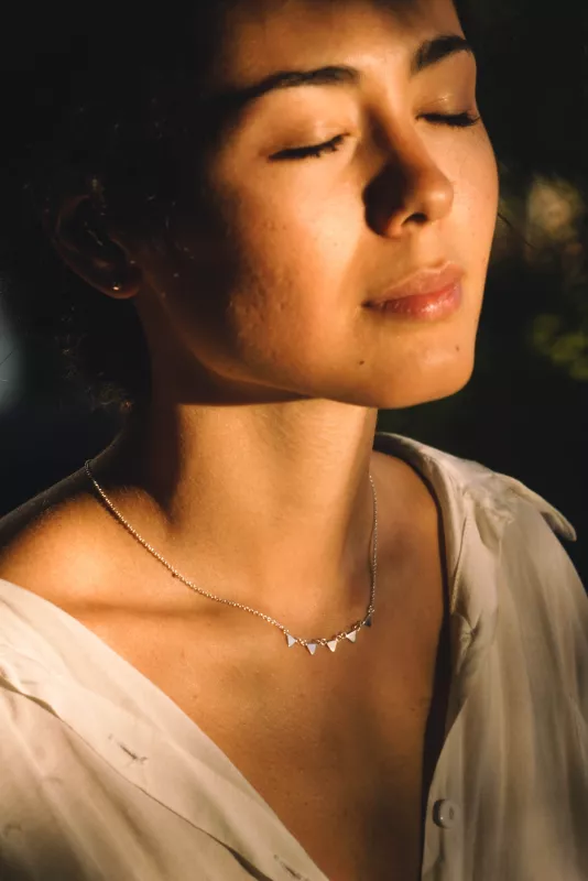 Young woman in sunlight with eyes closed, wearing delicate necklace, exudes tranquility and elegance.
