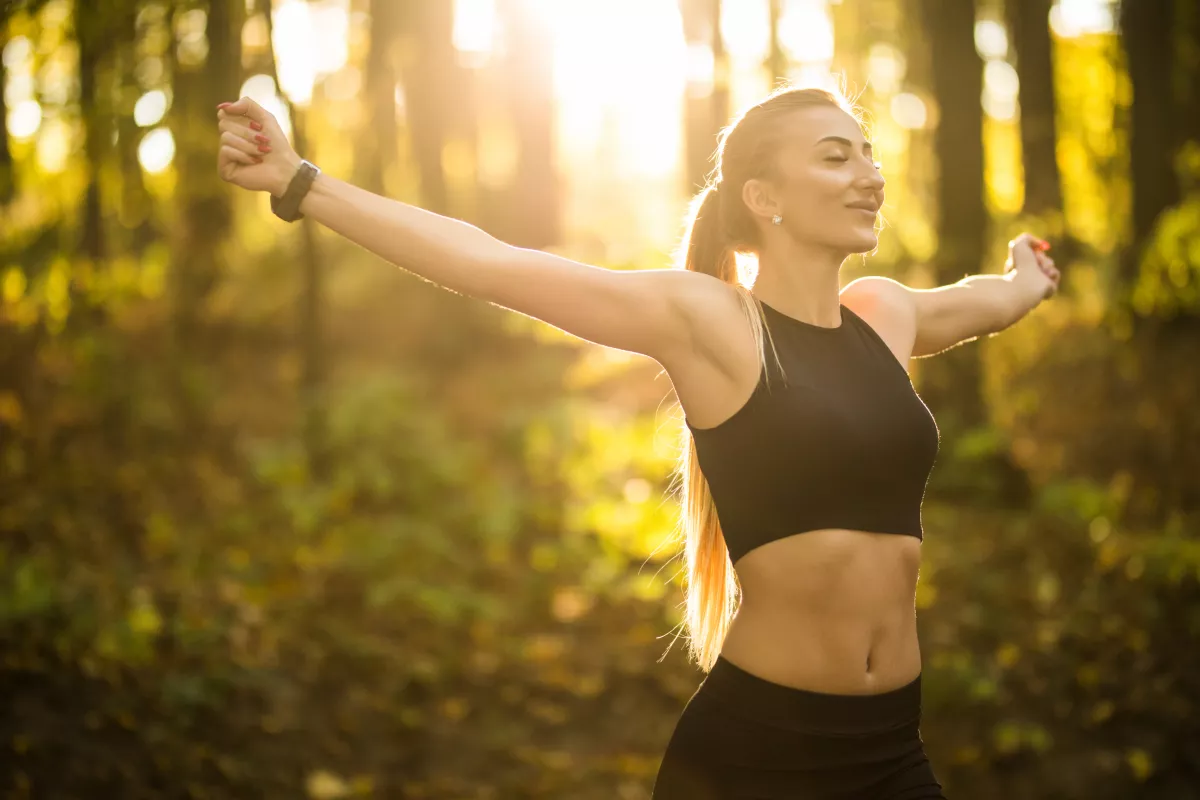 Woman meditating in forest, stretching in sunlight, showcasing health and wellness.