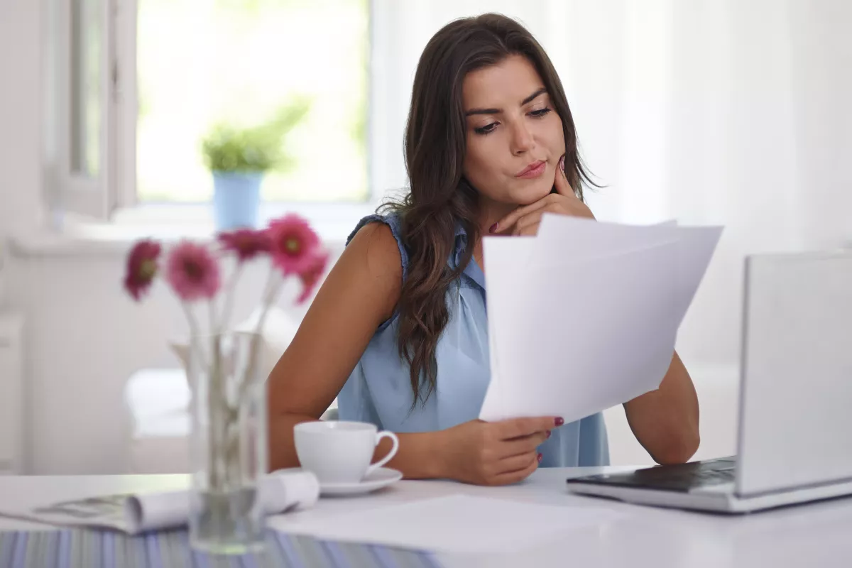 Woman contemplating papers at desk with laptop and flowers in a bright, serene workspace.