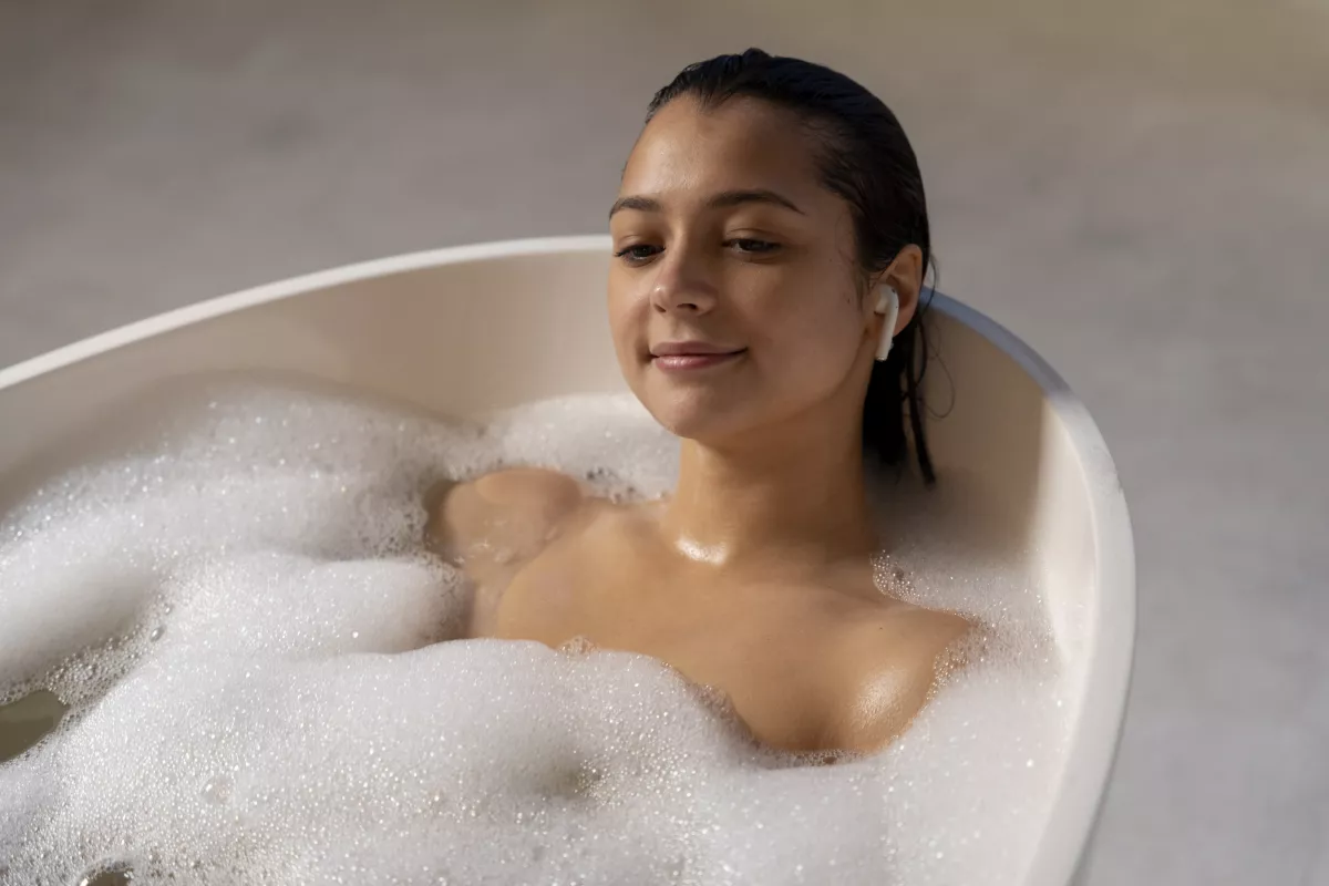 Woman in bubble bath with earbuds enjoys serene self-care moment with CBD bath bomb.