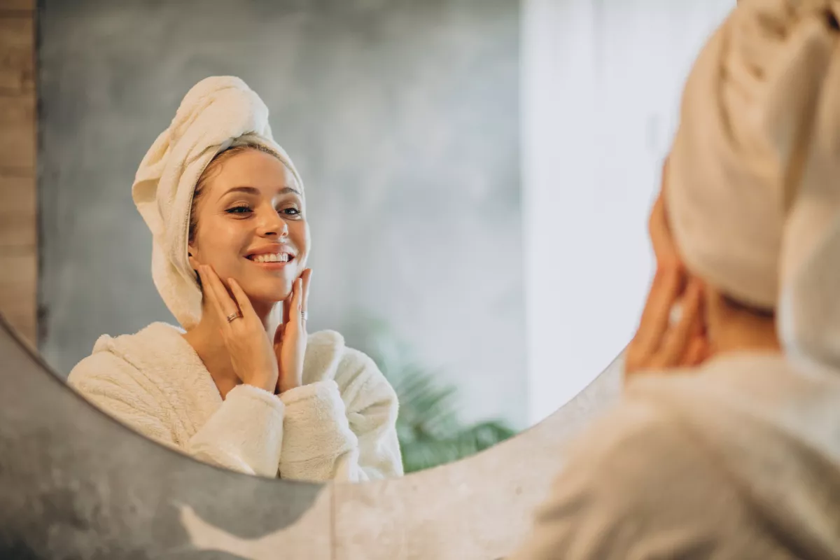 Woman in white robe enjoys skincare routine, smiling at reflection in round mirror with greenery background.