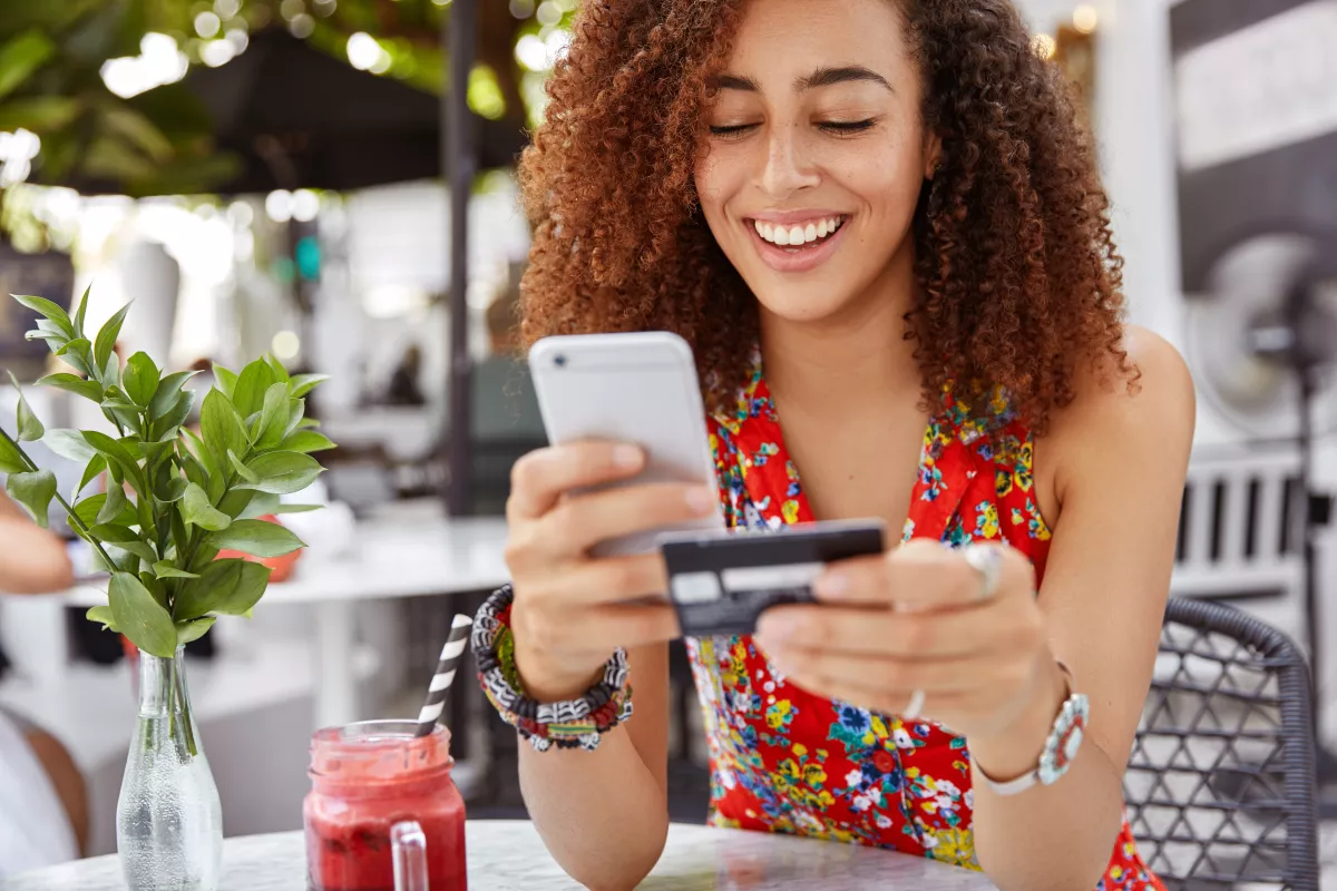 Woman using credit card and smartphone at sunny café surrounded by greenery.
