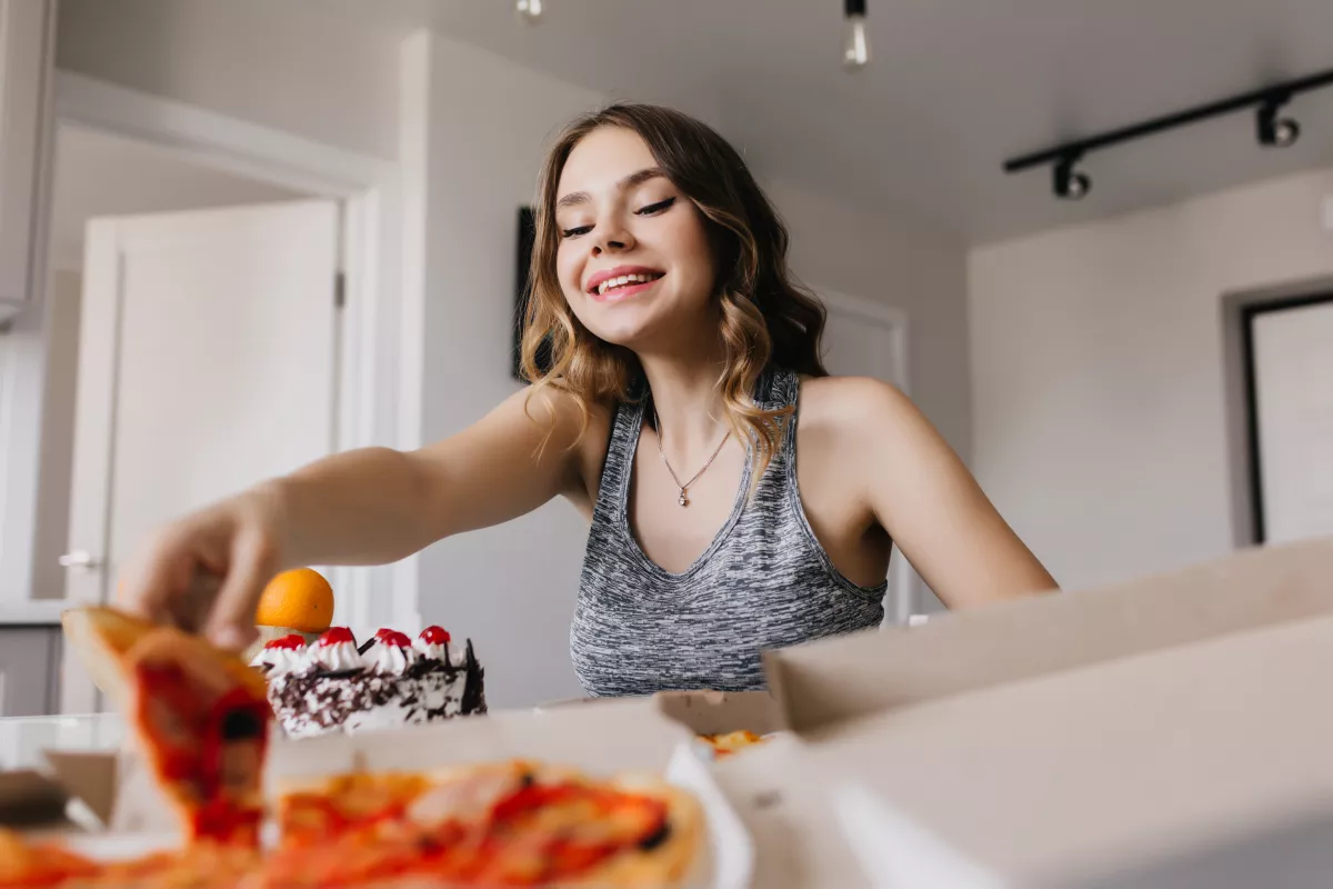 Woman in tank top smiling while grabbing pizza slice in a bright, stylish kitchen setting.