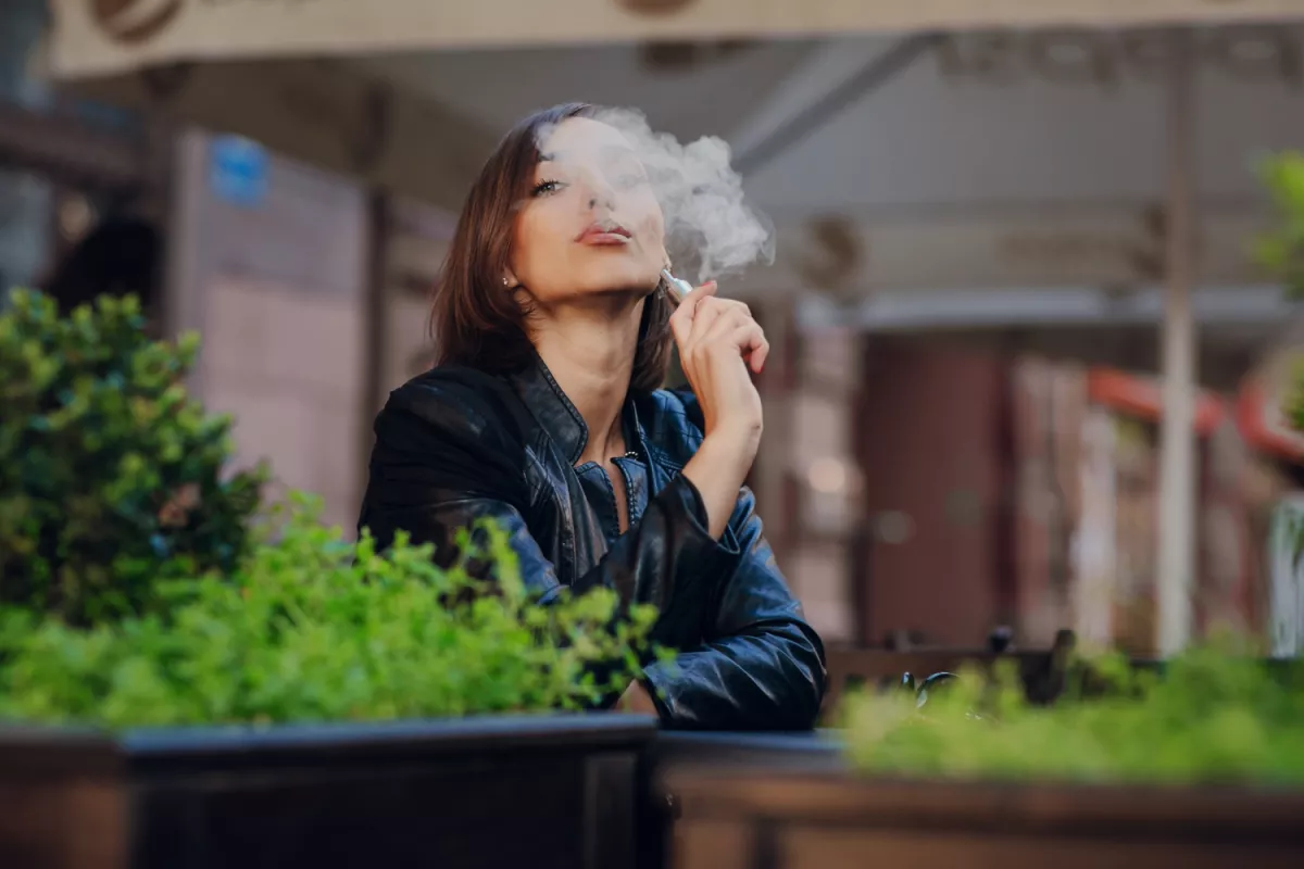 Woman in leather jacket smoking cigarette by green plants outdoors.