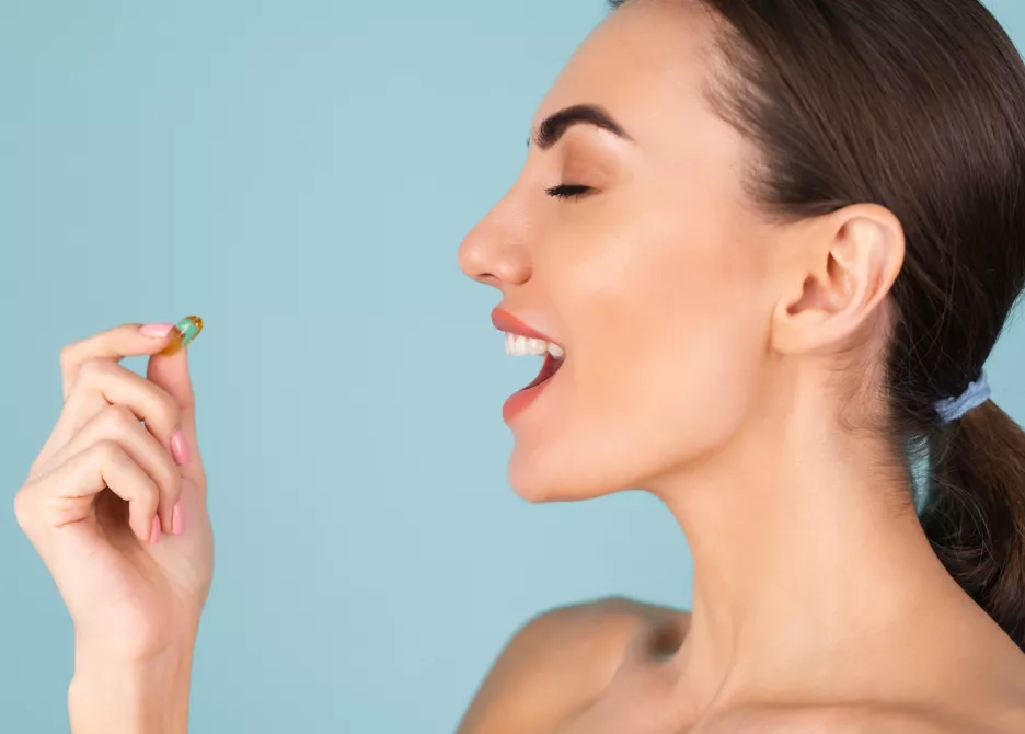 Woman with radiant skin ingests dietary supplement pill against light blue backdrop.