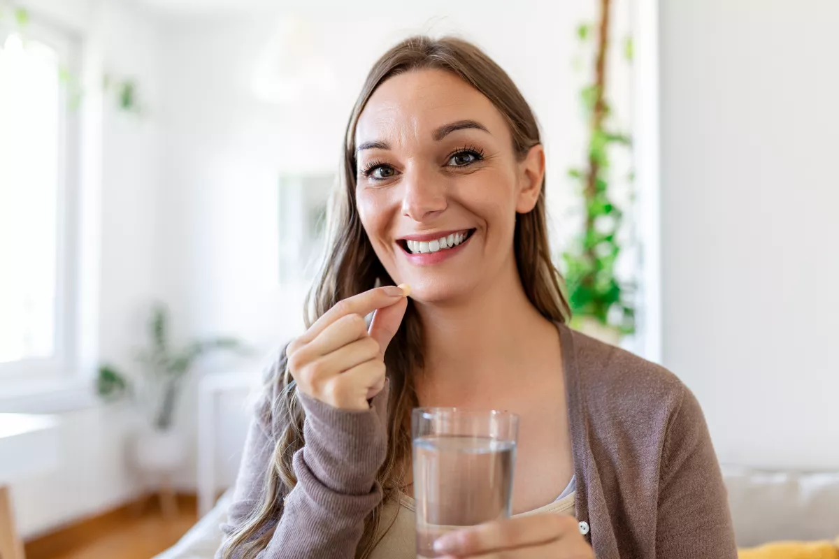 Woman smiling, holding water and CBD pill in cozy home setting.