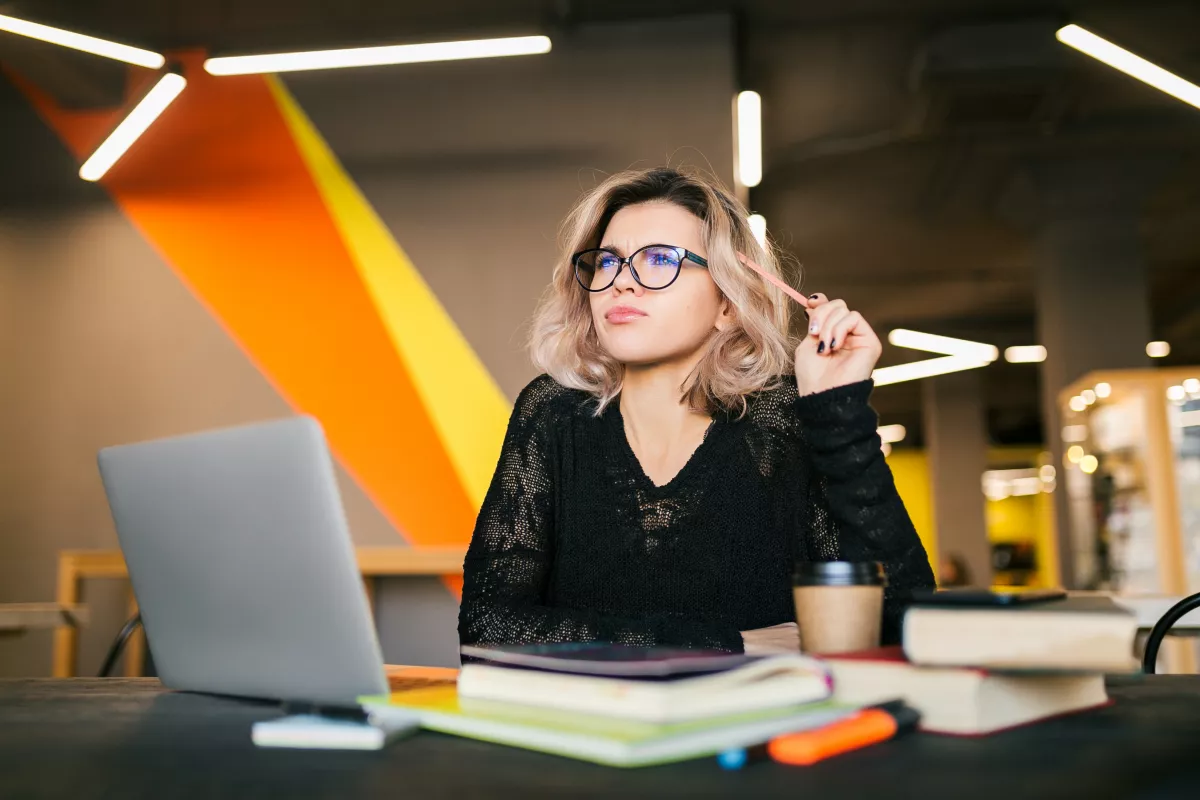 Woman in glasses deep in thought at a desk with laptop, notebook, and coffee cup.