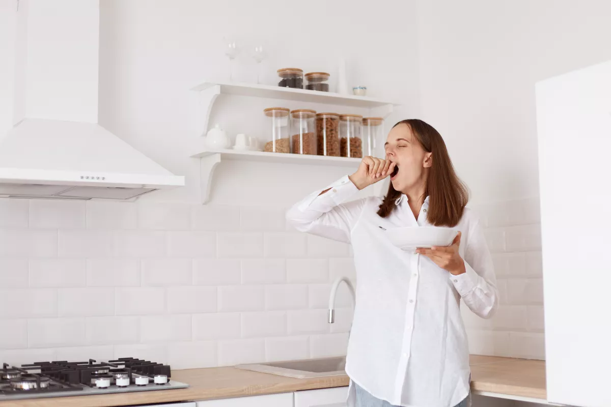 Woman yawning in sleek, modern kitchen with minimalist design and natural light.