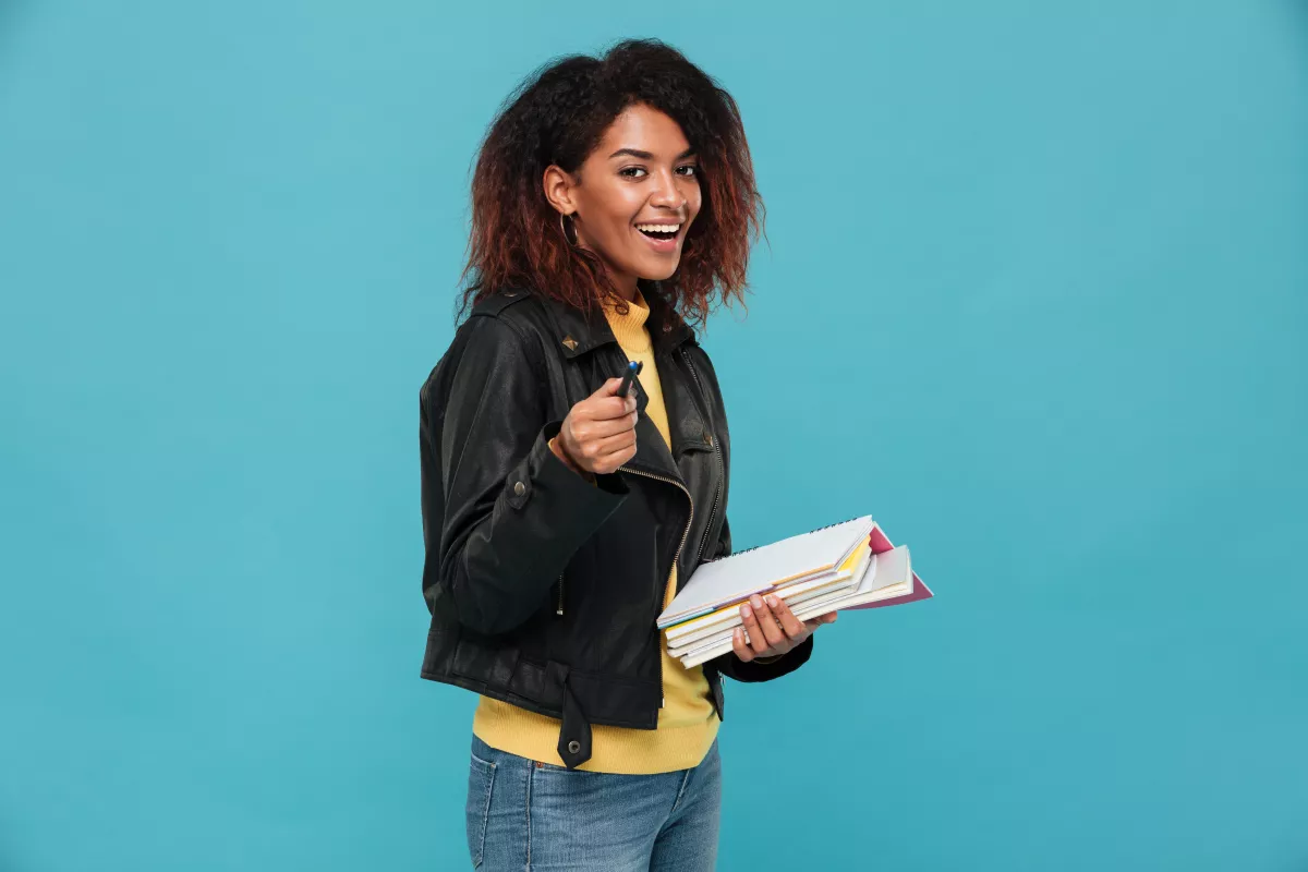 Happy student with books and pen against turquoise backdrop, prepared for class.