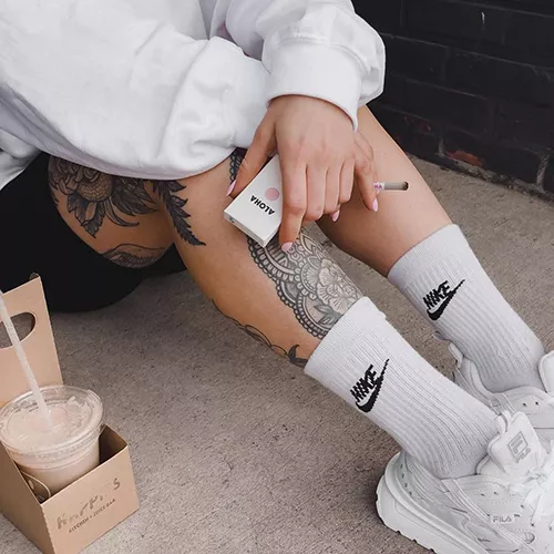 Urban street style: tattooed legs, white oversized sweater, Nike socks, Fila sneakers, cold coffee.