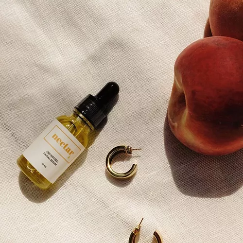 Serene arrangement of nectar oil, gold hoop earrings, and ripe peaches on linen fabric.
