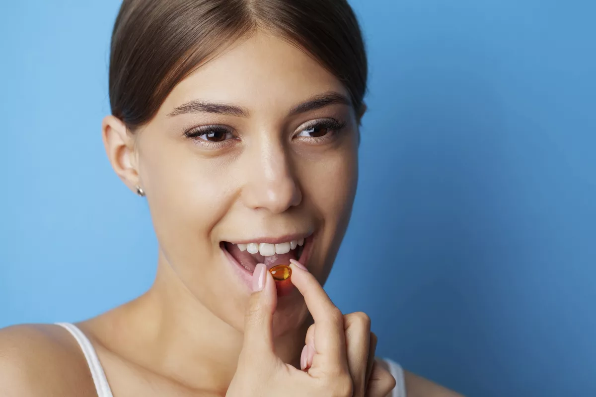 Woman smiling, taking amber CBD capsule for wellness against blue background.