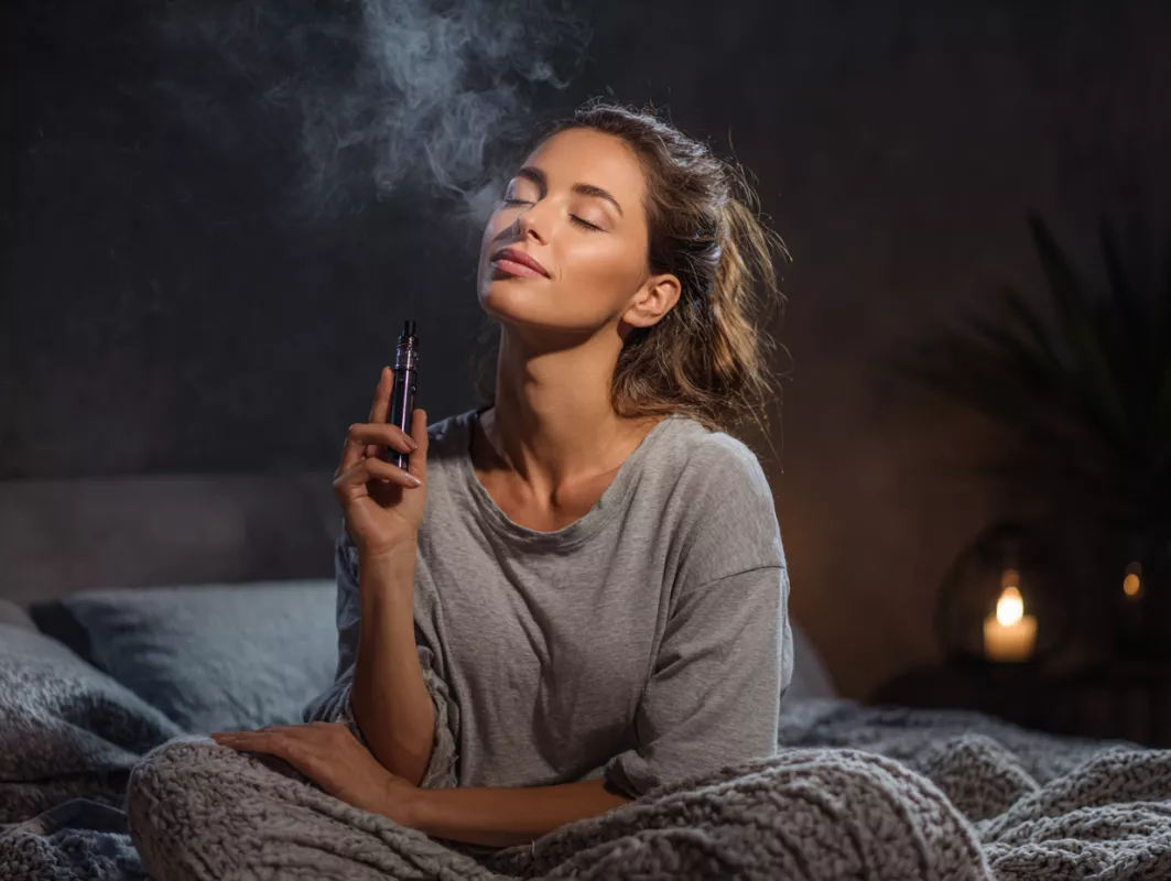 Woman relaxing in dimly lit room, vaping on bed, exuding cozy and peaceful vibes.