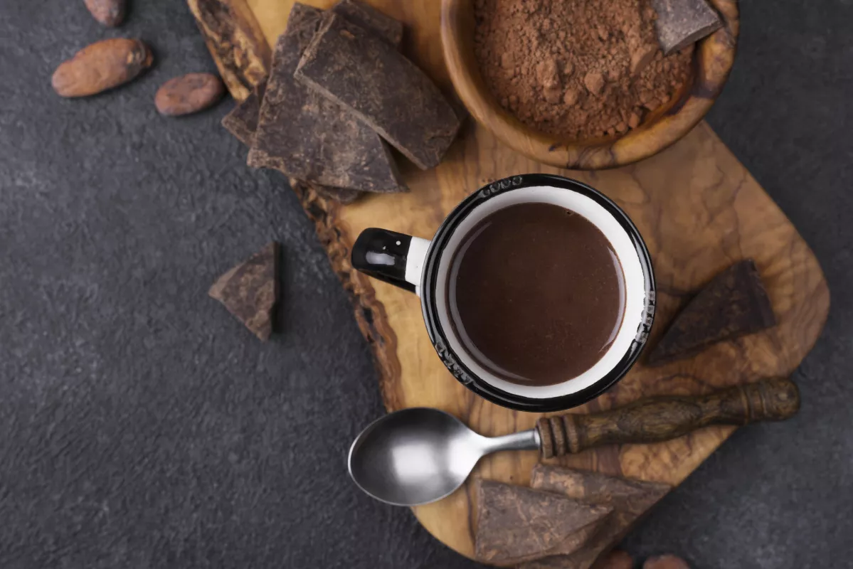 Rustic hot chocolate, cocoa powder, wooden spoon, and beans arrangement on dark background.