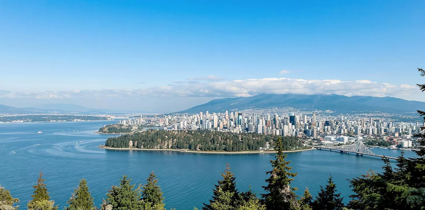 Cityscape with skyscrapers framed by lush forests, calm water, and majestic mountains.