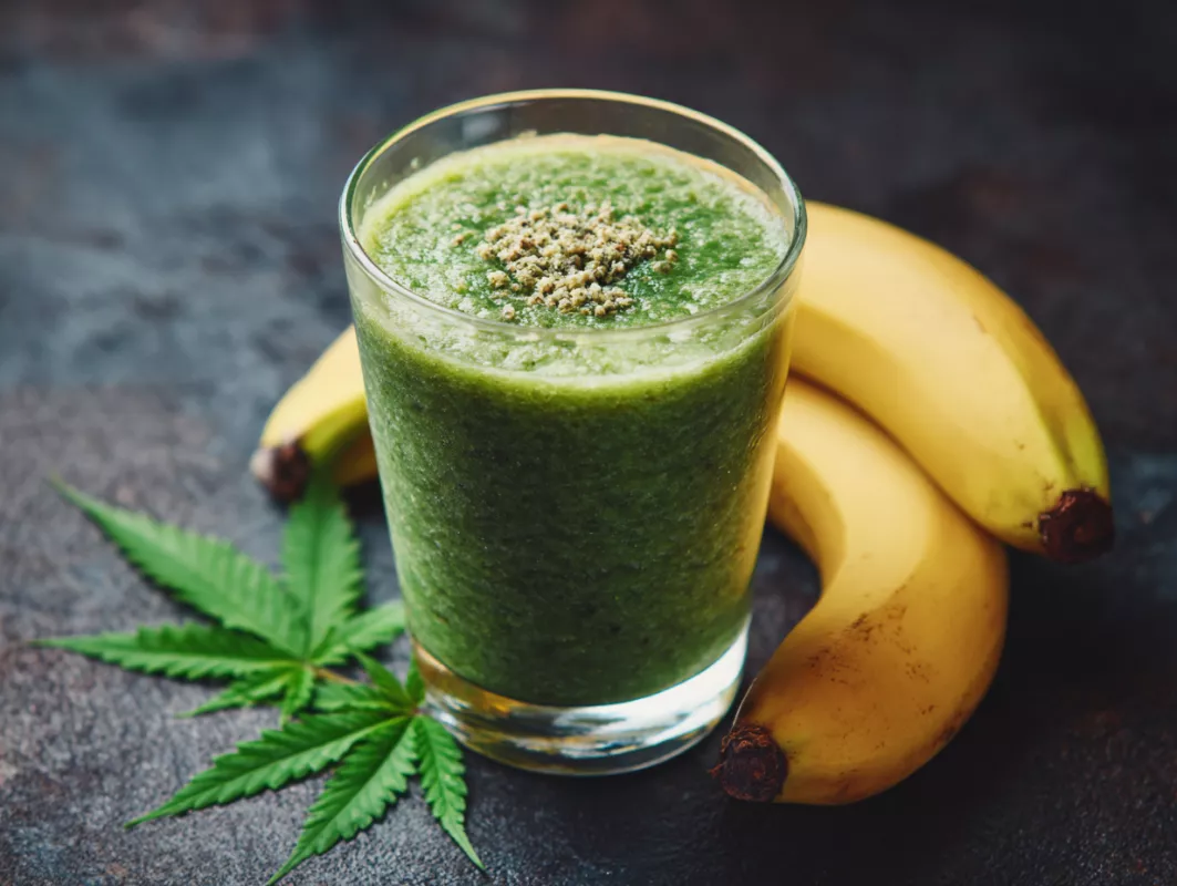 Glass of green smoothie with bananas and leafy garnish on a dark background.