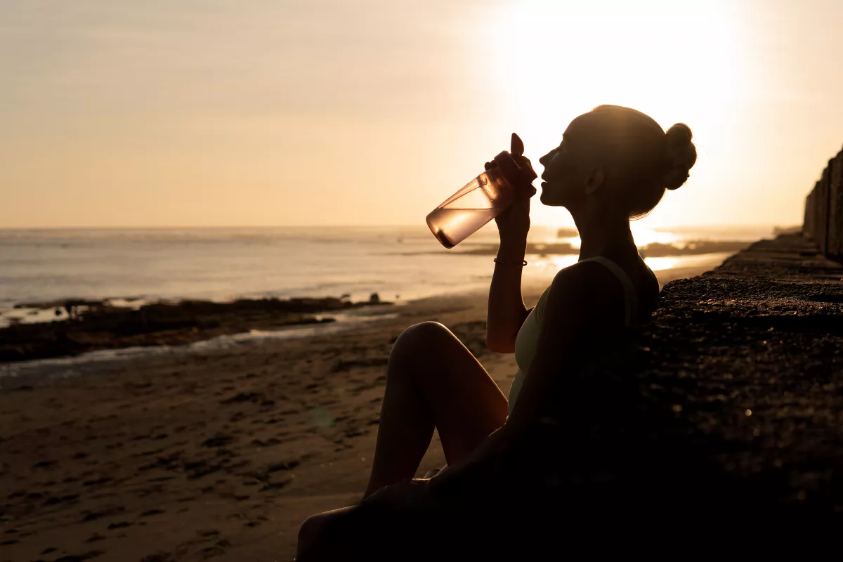Woman sipping CBD water on beach at sunset, silhouette against shimmering ocean.