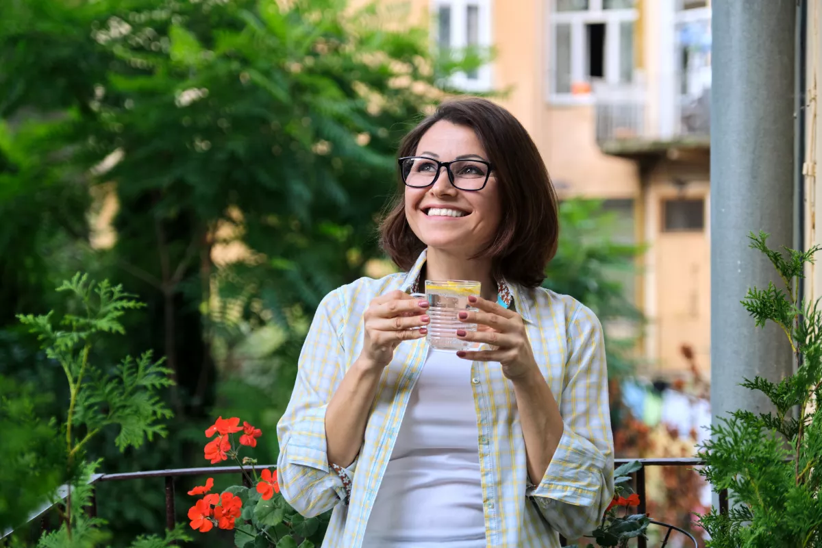 Woman sipping yellow drink on sunny urban balcony surrounded by plants and flowers.