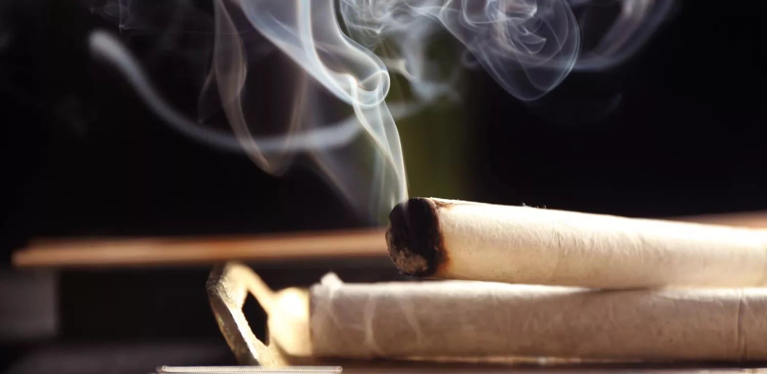 Glowing ember on hand-rolled cigarette with rising smoke against dark background.
