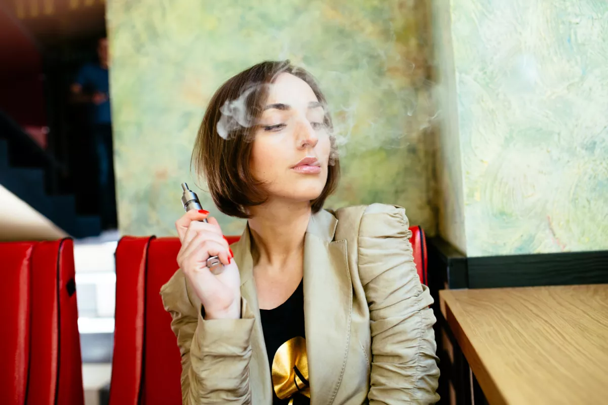 Person vaping Delta 8 cannabis e-cigarette in a chic cafe with colorful art backdrop.