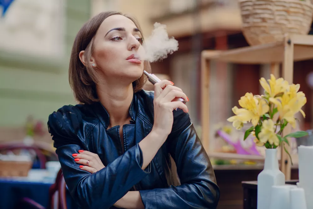 Woman in leather jacket enjoying a peaceful vape moment with yellow flowers at a cafe.