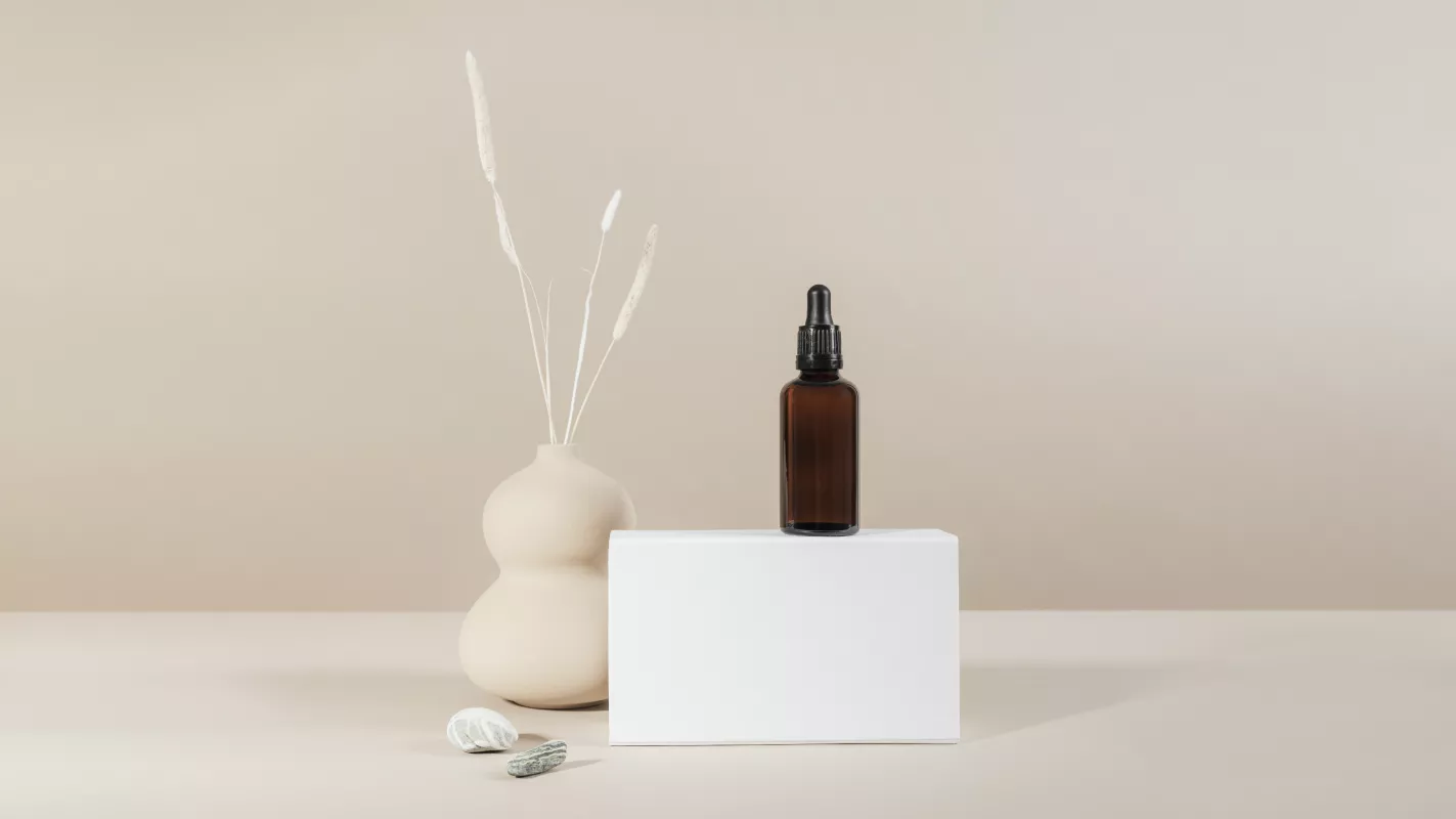 Amber glass dropper bottle on white pedestal with vase and botanical stems for essential oils.