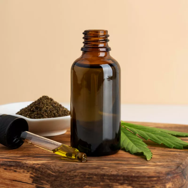 Amber bottle with cannabinoid oil, dropper, herbs, and cannabis leaves on wood.