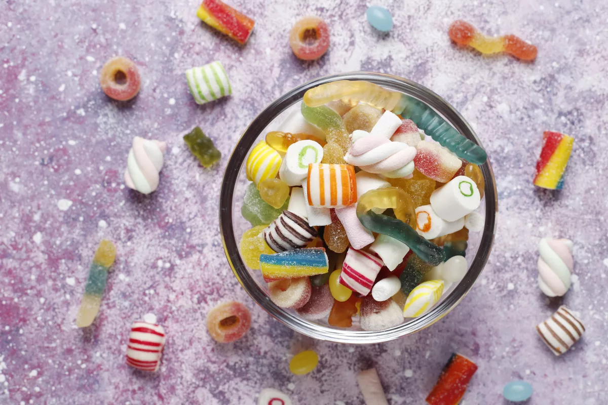 Colorful vegan candies with gummy worms and hard treats in glass bowl on purple background.