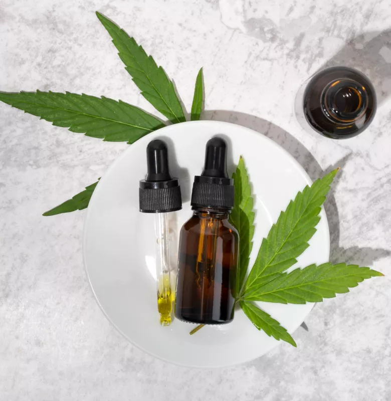 Amber dropper bottles with CBD oil on marble, surrounded by fresh cannabis leaves.