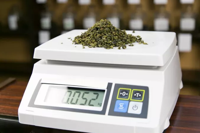 Accurate digital scale displaying 0.052 kg of green, leafy cannabis on wood with blurred jars behind.