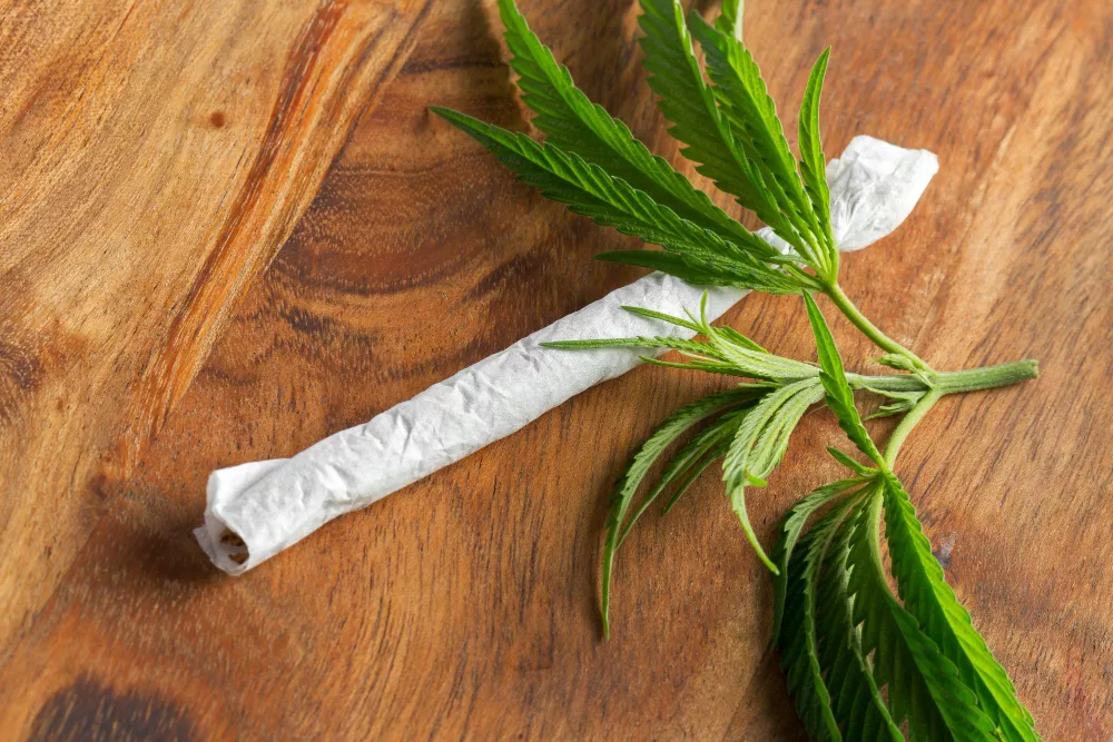 Hand-rolled cannabis joint with fresh leaves on a wooden surface.