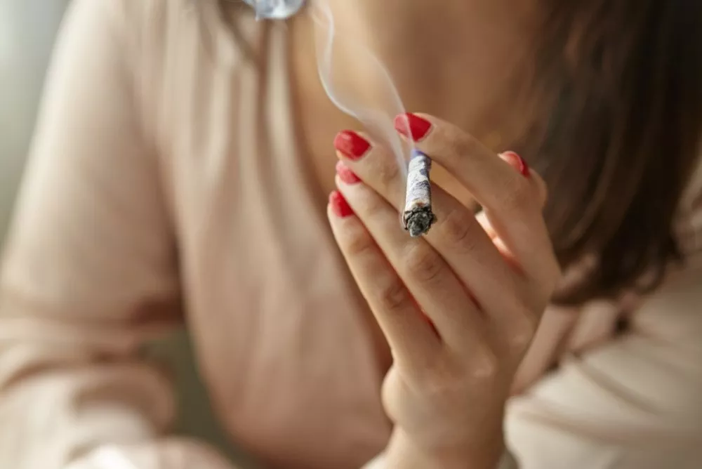 Adult with red manicure holding lit cigarette, smoke rising against soft background.