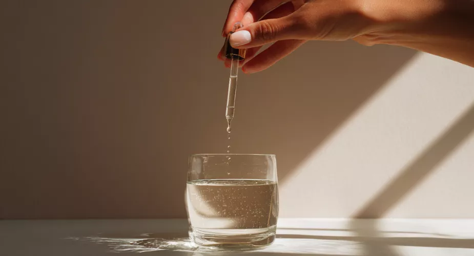 Hand holding a dropper above a brown glass bottle, against a soft yellow background.